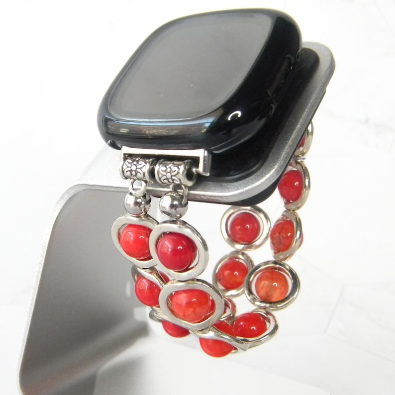 May include: A black smartwatch with a red beaded band. The band is made of small, round beads with silver accents.