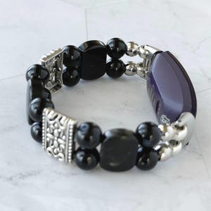 May include: A black beaded bracelet with silver-toned accents. The bracelet features a mix of round and square black beads, interspersed with silver beads and decorative silver clasps. A purple and silver component is also included.