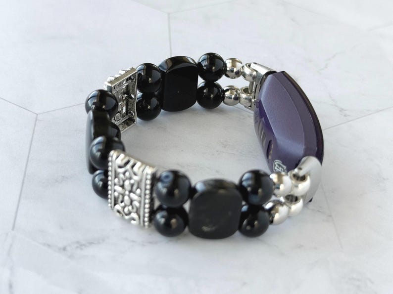 May include: A black beaded bracelet with silver-toned accents. The bracelet features a mix of round and square black beads, interspersed with silver beads and decorative silver clasps. A purple and silver component is also included.