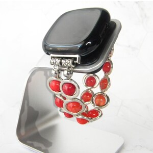 May include: A black smartwatch with a red beaded bracelet. The bracelet is made of small, round beads that are strung together on a silver chain. The bracelet is attached to the watch with a silver clasp.