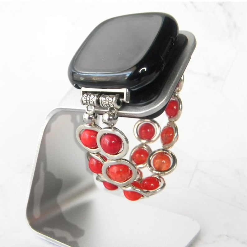 May include: A black smartwatch with a red beaded bracelet. The bracelet is made of small, round beads that are strung together on a silver chain. The bracelet is attached to the watch with a silver clasp.
