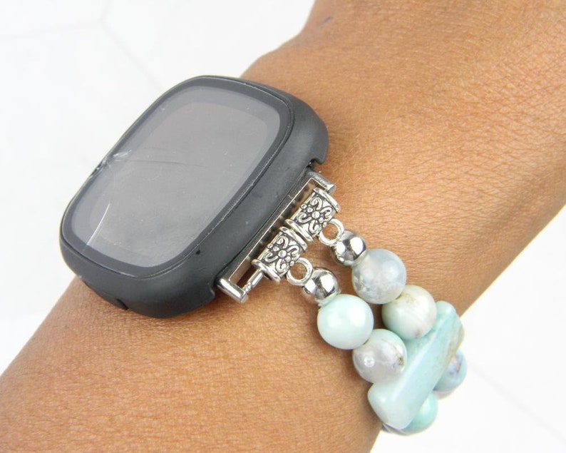 May include: A black smartwatch with a cracked screen is attached to a light blue beaded bracelet with silver accents.
