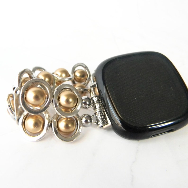 May include: A gold and silver beaded watch band with a black smartwatch. The band is made of small, round beads that are connected by silver rings.