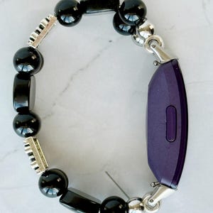 May include: A black beaded bracelet with a purple fitness tracker. The bracelet features a mix of round and rectangular black beads, interspersed with silver-colored metal accents. The fitness tracker is a deep purple color and is connected to the bracelet with silver-colored clasps.