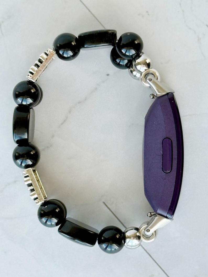 May include: A black beaded bracelet with a purple fitness tracker. The bracelet features a mix of round and rectangular black beads, interspersed with silver-colored metal accents. The fitness tracker is a deep purple color and is connected to the bracelet with silver-colored clasps.