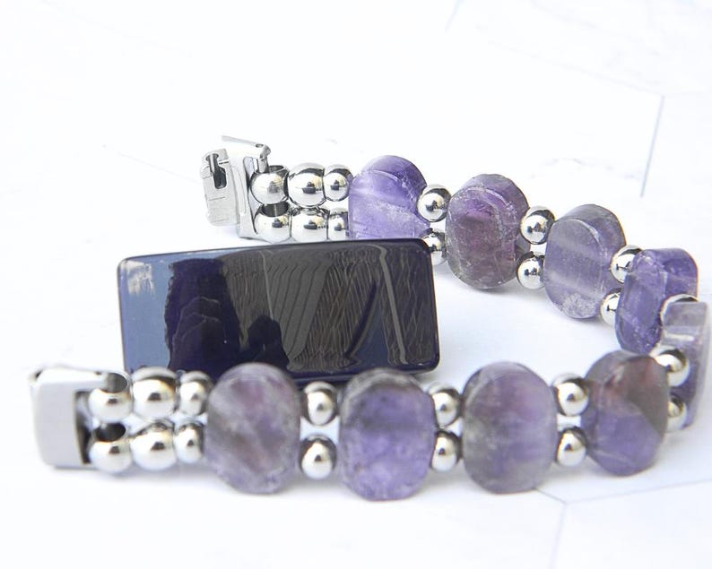 Puede incluir: Un brazalete con piedras preciosas ovales de color p&uacute;rpura y peque&ntilde;as cuentas plateadas. El brazalete tiene un cierre plateado y se muestra junto a un objeto rectangular azul oscuro. Las piedras preciosas tienen tonos p&uacute;rpuras claros y oscuros.