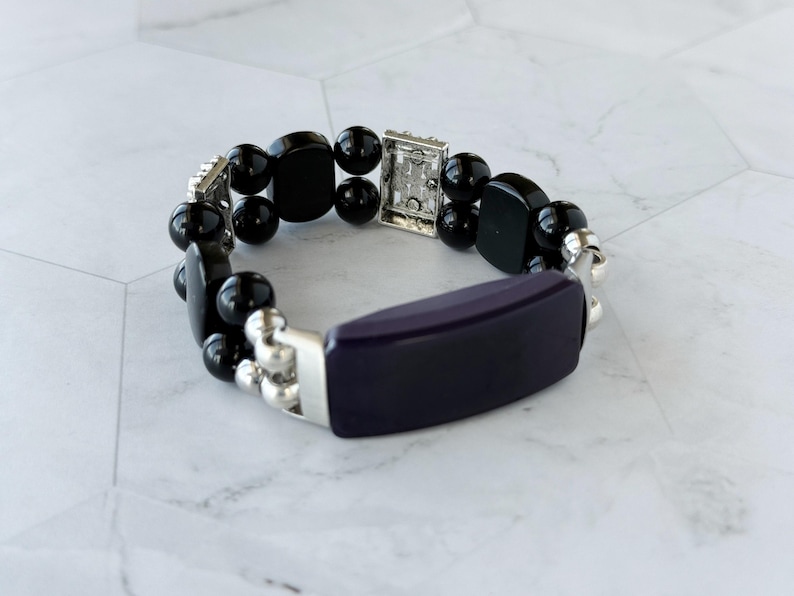 May include: A beaded bracelet featuring a dark purple rectangular center piece and black, rounded beads. The bracelet has silver-toned metal accents and connectors. The jewelry is displayed on a white and gray surface.