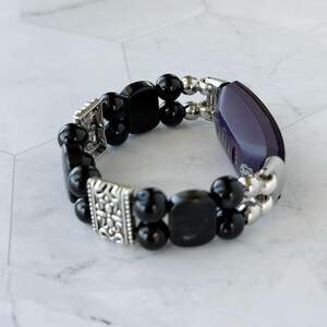 May include: A beaded bracelet featuring black, rounded beads and silver-toned accents. The bracelet includes a dark purple, oval-shaped element and decorative silver metal pieces. The design is a mix of classic and modern styles.