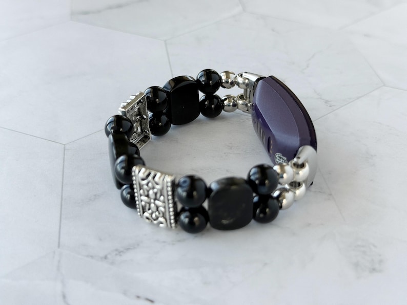 May include: A beaded bracelet featuring black, rounded beads and silver-toned accents. The bracelet includes a dark purple, oval-shaped element and decorative silver metal pieces. The design is a mix of classic and modern styles.