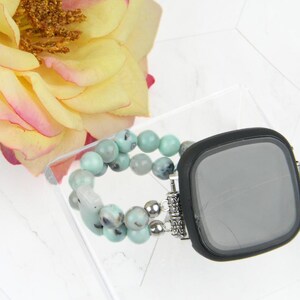 May include: A black smartwatch case with a turquoise and silver beaded band. The case has a round screen and a silver metal frame. The band is made of small, round beads that are turquoise and silver in color.