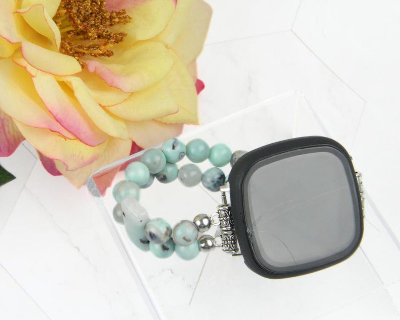 May include: A black smartwatch case with a turquoise and silver beaded band. The case has a round screen and a silver metal frame. The band is made of small, round beads that are turquoise and silver in color.