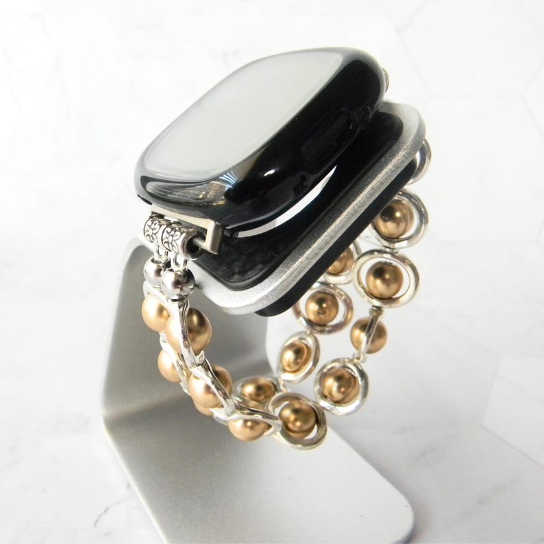 May include: A black smartwatch with a gold and silver beaded band. The watch is on a silver stand.