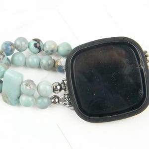 May include: A black smartwatch with a light blue and white beaded band. The band is made of multiple strands of beads and has a silver clasp.