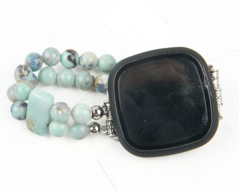 May include: A black smartwatch with a light blue and white beaded band. The band is made of multiple strands of beads and has a silver clasp.