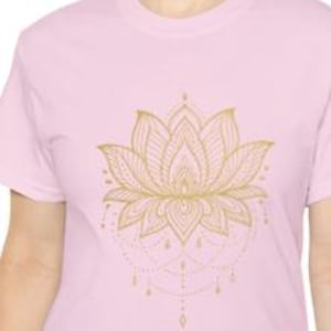 May include: A pale pink t-shirt with a gold lotus flower design. The detailed floral pattern includes fine lines and a dotted mandala-style base. The shirt has a crew neck and short sleeves.