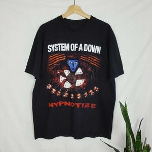 May include: Black t-shirt featuring the band name "SYSTEM OF A DOWN" in white block letters above a graphic. The graphic includes a blue-faced figure, clock-like design, and the word "HYPNOTIZE" in red.