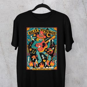 May include: Black t-shirt featuring a colorful graphic of a musician playing a guitar. The design includes the words "Billy Strings" and the date and location of a concert. The artwork has a psychedelic, vintage concert poster style.