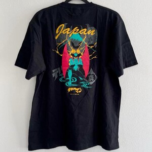 May include: Black t-shirt with a graphic design on the back featuring the word "Japan" in yellow and teal. The graphic includes a figure with red wings and a teal and yellow color scheme. The Japanese text is also visible.