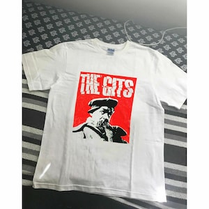 May include: White t-shirt with a red square graphic featuring the text "THE GITS" above a black and white portrait of a man wearing a hat. The shirt is made of a soft material.