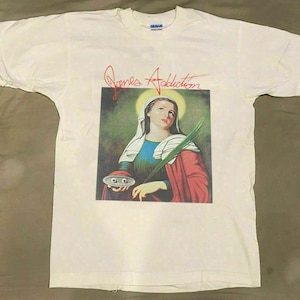 May include: White t-shirt with a graphic of a woman with a halo, holding a palm frond and a plate with two eyes. The text "Jane's Addiction" is written in red cursive above the graphic. The shirt is a Gildan brand.