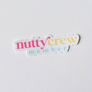 May include: A white sticker with the words "nutty crew member" in pink, yellow, and light blue. The sticker has a rounded rectangular shape and is placed on a white background.