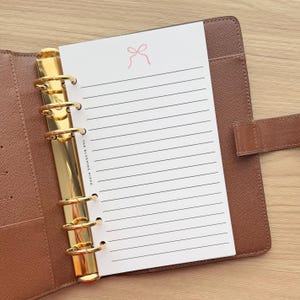 May include: A brown textured leather planner with a gold-toned six-ring metal binder mechanism. Inside is a white lined paper insert featuring a small pink bow icon at the top and the text THE BLUSHING HIPPO printed vertically along the left margin.