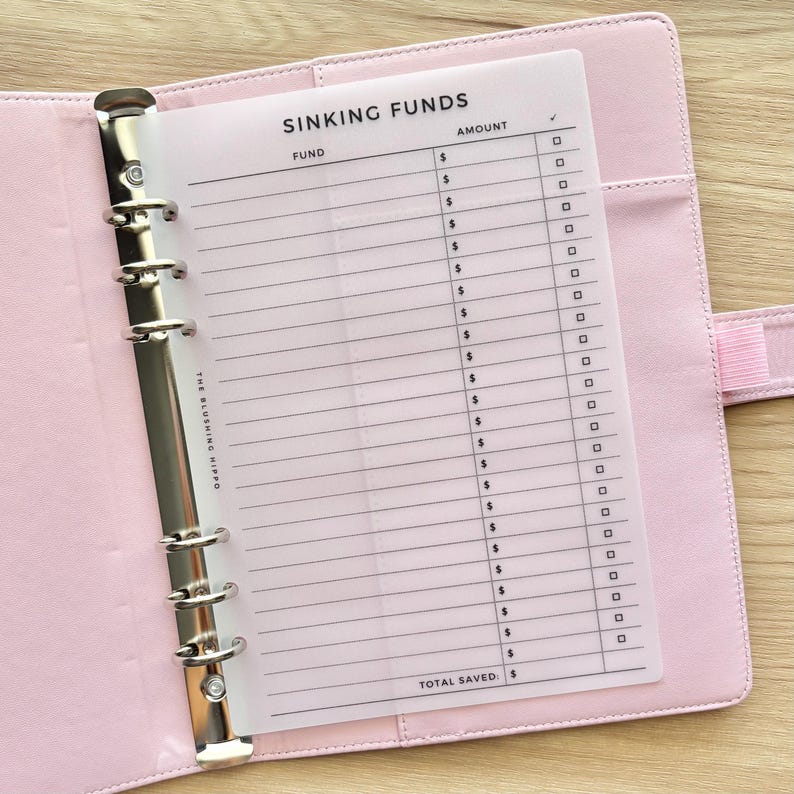 May include: A light pink binder featuring a translucent insert page titled SINKING FUNDS. The page includes columns for Fund, Amount, and a checkbox, with the text THE BLUSHING HIPPO printed vertically along the left side and TOTAL SAVED at the bottom.