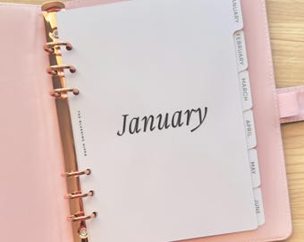 May include: A light pink personal planner with a rose gold six-ring metal binder mechanism. The white interior page features the word January in black script, with the text The Blushing Hippo printed vertically and month tabs for January through June.