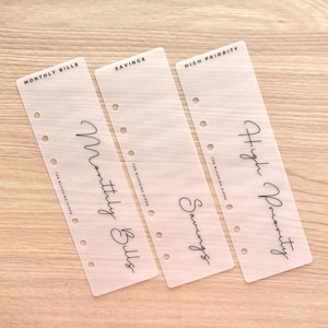 May include: Three translucent planner inserts with black text. The inserts are labeled "Monthly Bills", "Savings", and "High Priority". Each insert has pre-punched holes for binding. The brand name "The Blushing Hippo" is printed at the bottom of each insert.