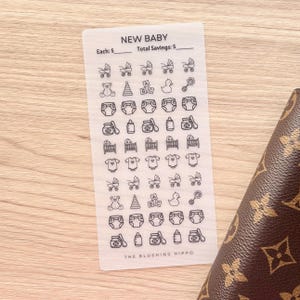 May include: A white sticker sheet with the text "NEW BABY" and various baby-themed illustrations. The sheet includes spaces to track savings. A brown patterned wallet is partially visible in the image.