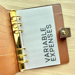 May include: A brown leather-textured planner with a gold-toned six-ring metal binder mechanism. A translucent divider page is inserted, featuring the black printed text "VARIABLE EXPENSES" and the small brand name "THE BLUSHING HIPPO" vertically.