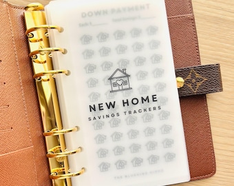 May include: A white plastic sheet with a black and white house icon and the text "NEW HOME SAVINGS TRACKERS" printed on it. The sheet is designed to be used as a savings tracker for a new home purchase. The sheet is placed in a brown leather planner.