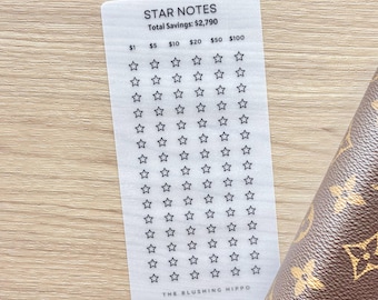 May include: A white Star Notes savings tracker card with a grid of stars, with the text "STAR NOTES" and "Total Savings: $2,790". The card has denominations of $1, $5, $10, $20, $50, and $100. The Blushing Hippo logo is at the bottom. A brown patterned wallet is in the background.