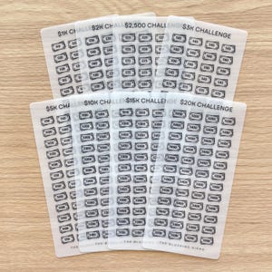 May include: A collection of clear, rectangular challenge cards with various dollar amounts printed on them. The cards are labeled with challenges ranging from $1K to $20K. Each card has multiple boxes with dollar amounts to be checked off.
