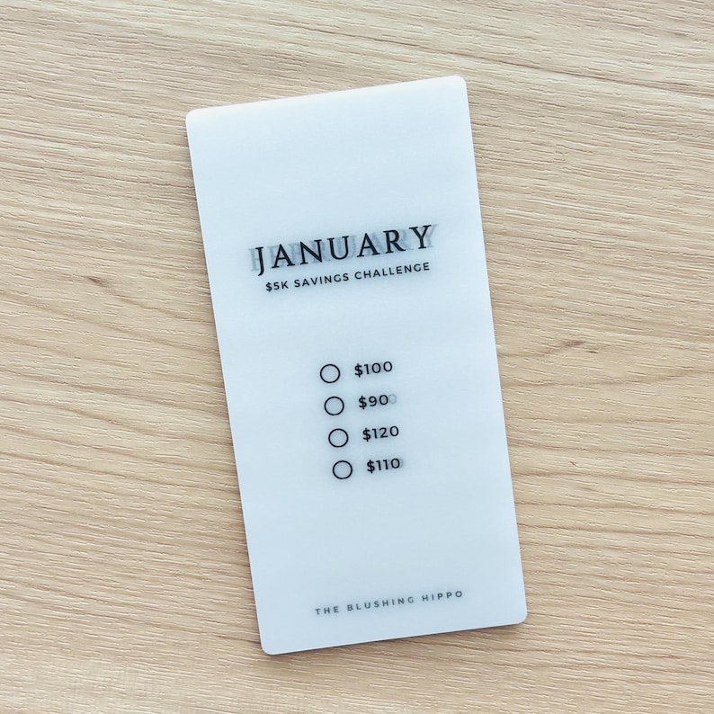 May include: White card with the text "JANUARY $5K SAVINGS CHALLENGE" and a list of dollar amounts to save each month: $100, $90, $120, $110. The bottom of the card reads "THE BLUSHING HIPPO".