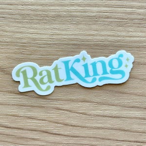 May include: A white sticker with the words "RatKing" in a retro font. The word "Rat" is in light green, and "King" is in turquoise blue with star accents. The sticker is on a light brown wooden surface.