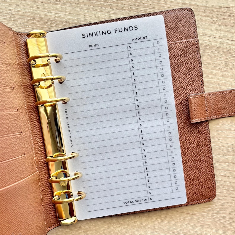 May include: A brown leather-textured binder with a gold-toned six-ring mechanism holding a white translucent insert. The insert features the header SINKING FUNDS, columns for FUND, AMOUNT, and checkboxes, with the text THE BLUSHING HIPPO printed vertically.