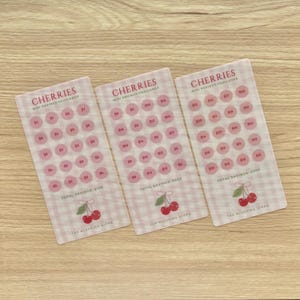 May include: Three pink and white patterned mini savings challenge cards with the word "CHERRIES" at the top. Each card has circles with dollar amounts and a cherry graphic. The total savings amounts are $100, $200, and $300.