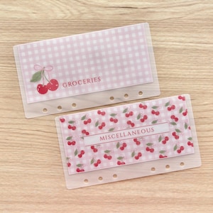 May include: Two translucent plastic binder inserts with six-hole punches. One features a pink and white gingham pattern with a cherry illustration and the text GROCERIES. The other has a repeating cherry pattern and the text MISCELLANEOUS.