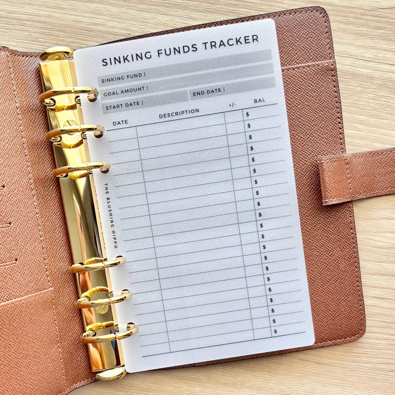 Op de afbeelding: Een bruine, getextureerde lederen map met een gouden zes-ringsmechanisme met een wit Sinking Funds Tracker-inlegvel. De pagina bevat velden voor Sinking Fund, Goal Amount, Start Date, End Date en een tabel met kolommen voor Date, Description, +/- en Bal.
