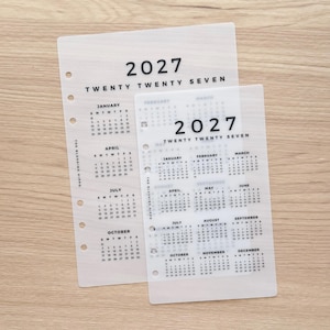 May include: Two translucent, frosted plastic planner inserts featuring the 2027 calendar year. Each sheet displays the year 2027 in bold black text, followed by monthly grids with days of the week. The text The Blushing Hippo is printed vertically.