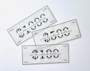 May include: Three rectangular translucent acrylic cards resembling currency. The cards feature black printed text and numbers: $1000 with one thousand dollars, $500 with five hundred dollars, and $100 with one hundred dollars in a script font.