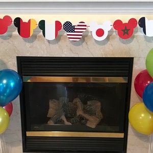 May include: A fireplace mantel decorated with a Mickey Mouse themed banner featuring flags from different countries. A cluster of balloons in red, blue, yellow, and green are arranged in front of the fireplace.