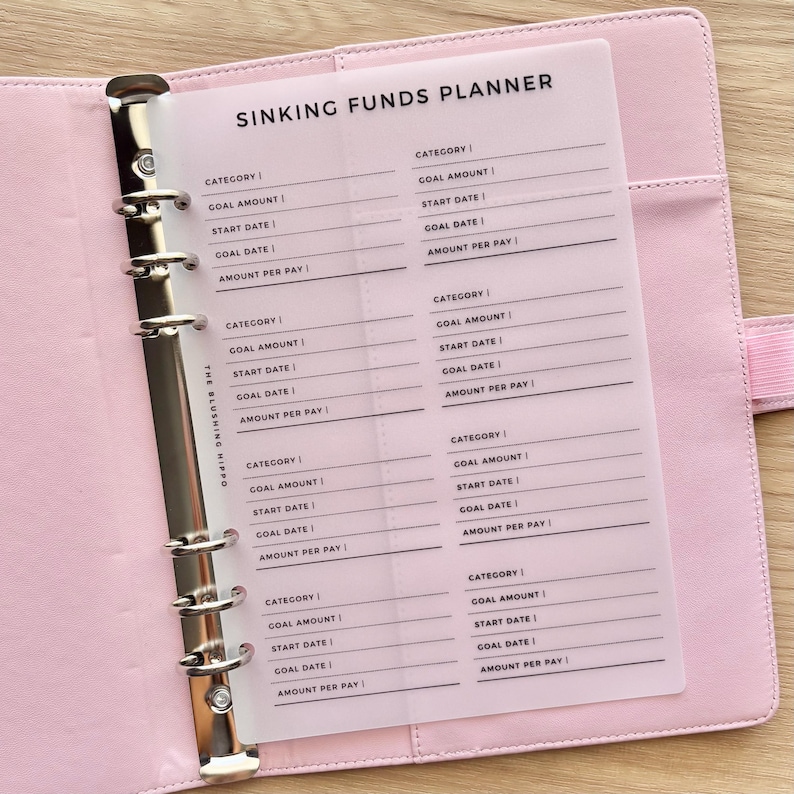 May include: A light pink six-ring binder containing a translucent plastic insert titled SINKING FUNDS PLANNER. The insert features printed black text with fields for category, goal amount, start date, goal date, and amount per pay for tracking.