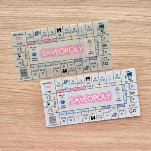 May include: Two rectangular game boards with the text "SAVEOPOLY" in pink. The boards have spaces labeled "Parking," "Free," "In Jail," and "Go." The game boards are on a light brown wooden surface.