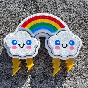 May include: A white keychain with a rainbow and two clouds with smiling faces and lightning bolts. The clouds have blue eyes and pink cheeks.