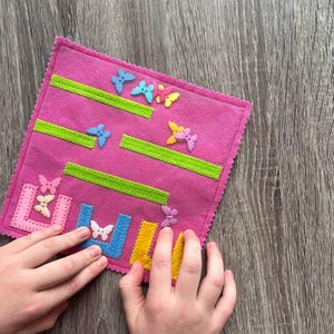 May include: A pink felt activity board with green stripes and colorful butterfly embellishments. The board has a stitched border and includes felt shapes in blue, yellow, and pink. A child's hands are interacting with the board.