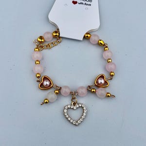Könnte beinhalten: Ein rosa und goldenes Perlenarmband mit einem großen Herz-Anhänger. Der Anhänger ist aus goldenem Metall und hat ein kleineres Herz im Inneren. Das Armband ist handgefertigt mit Liebe.