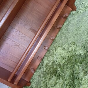 Puede incluir: Un estante de pared de madera de color marr&oacute;n medio que cuenta con una barra horizontal y una fila de seis peque&ntilde;os ganchos cil&iacute;ndricos que sobresalen. La unidad tiene un panel trasero plano con veta de madera visible y soportes laterales curvos.