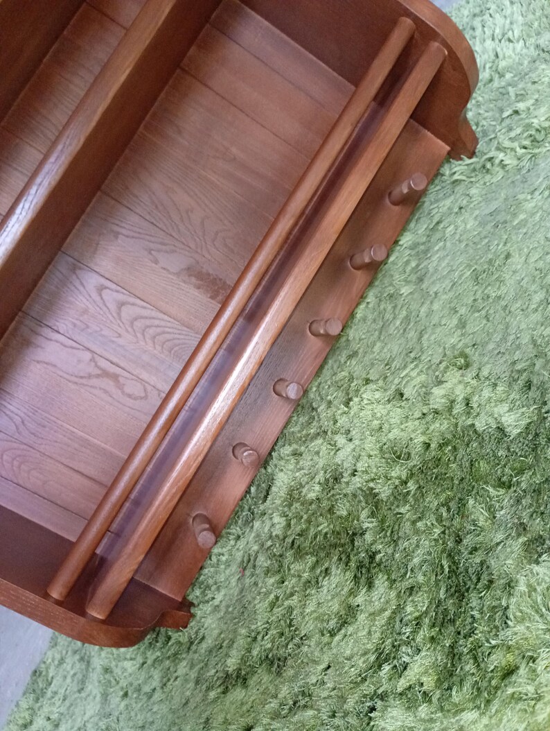 Puede incluir: Un estante de pared de madera de color marr&oacute;n medio que cuenta con una barra horizontal y una fila de seis peque&ntilde;os ganchos cil&iacute;ndricos que sobresalen. La unidad tiene un panel trasero plano con veta de madera visible y soportes laterales curvos.